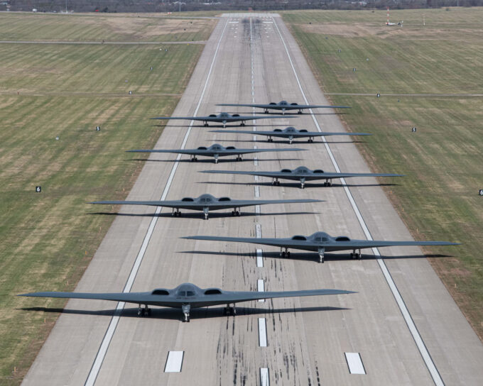 A imagem mostra uma pista longa com várias fileiras de aeronaves militares estacionadas nela. As aeronaves estão organizadas em uma linha arrumada e estão voltadas para o lado direito da imagem. A pista é cercada por campos verdes e existem alguns edifícios visíveis ao fundo. O céu é azul e o tempo parece estar ensolarado e claro. A imagem parece ter sido tirada de um ângulo alto, olhando para a pista.