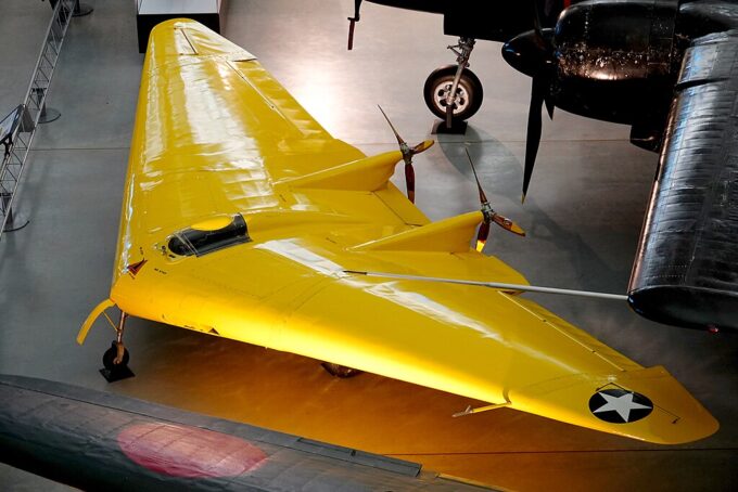 A imagem mostra um jato de caça amarelo estacionado em um hangar. O jato tem um nariz pontudo e duas hélices de ambos os lados. O corpo do jato é pintado em uma cor amarela brilhante com uma estrela preta na cauda. As asas também são pintadas na mesma cor amarela que o corpo. No fundo, existem outras aeronaves e uma escada encostada na parede. O hangar parece estar bem iluminado com luz natural chegando das janelas.