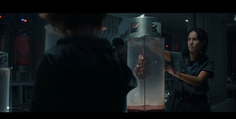 A imagem mostra uma cena de um filme ou programa de TV. Ele mostra duas pessoas, um homem e uma mulher, de pé em uma sala pouco iluminada com várias máquinas e equipamentos ao fundo. O homem está no lado esquerdo da imagem, vestindo uma jaqueta preta, e a mulher do lado direito está em pé na frente de um grande recipiente de vidro com uma tampa de metal. O recipiente parece ser preenchido com um líquido vermelho, possivelmente sangue ou sangue, e há uma pequena quantidade de sangue dentro. A mulher está segurando a tampa do recipiente com as duas mãos e está olhando para o homem com uma expressão séria em seu rosto. Parece que eles estão discutindo algo importante.