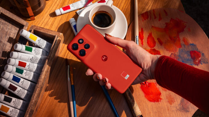 Moto Edge 50 Neo sendo segurado por uma mão estendida sobre uma mesa com café, canetas e pincéis de pintura