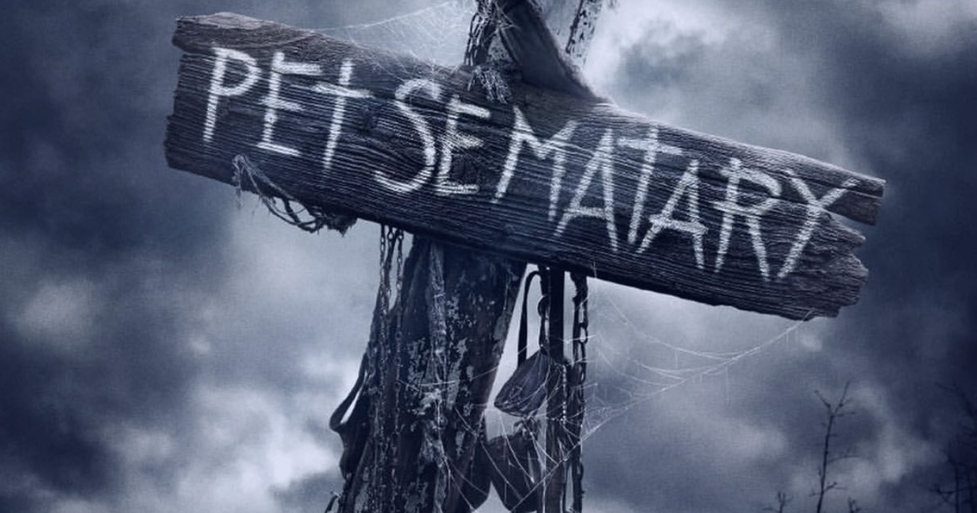 A imagem é uma fotografia em preto e branco de um cruzamento de madeira com as palavras "PET SEMATARY" escrito nela em giz branco. A cruz está presa a um tronco de árvore com uma teia de aranha pendurada nela. O fundo é escuro e nublado, com árvores nuas visíveis à distância. O humor geral da imagem é estranho e antecipado.