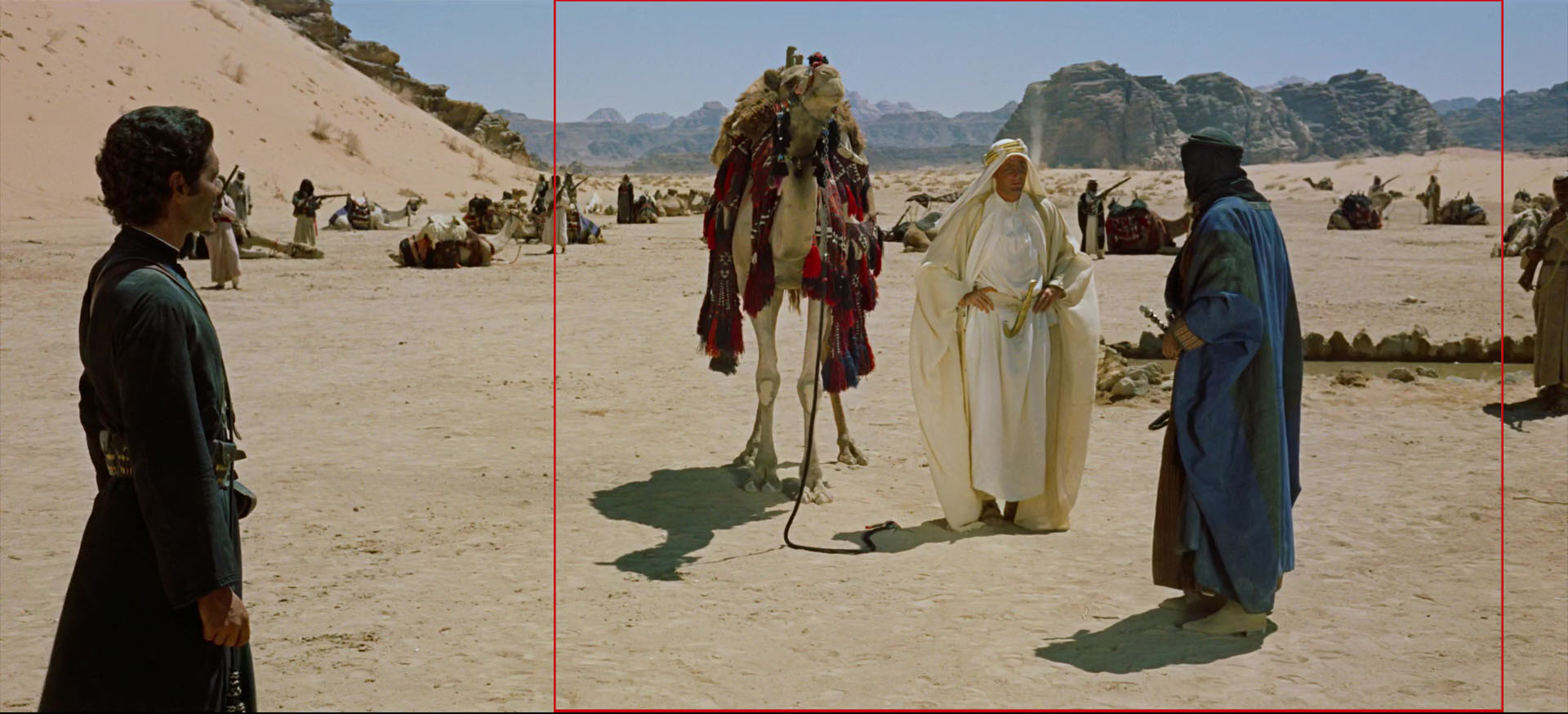 A imagem é uma colagem de três fotografias de uma cena do deserto. A primeira fotografia à esquerda mostra um homem em pé em frente a um camelo no deserto. O homem está usando uma túnica preta e parece estar olhando para o camelo com uma expressão séria no rosto. A segunda fotografia no meio mostra uma mulher vestindo uma túnica branca e um lenço na cabeça, parado ao lado do camelo. A terceira fotografia mostra um grupo de pessoas em segundo plano, algumas das quais estão sentadas e outras estão de pé. O fundo mostra uma vasta paisagem do deserto com montanhas à distância. O céu é azul e o humor geral da imagem é sombrio.