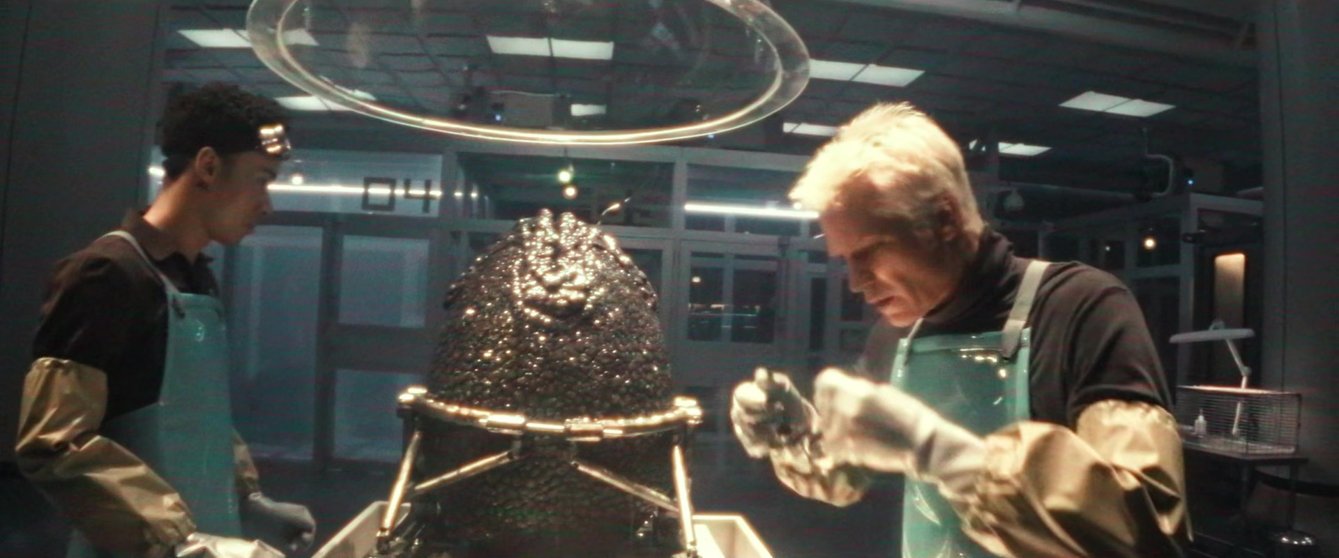 A imagem mostra dois homens em laboratório. Ambos estão usando aventais azuis e parecem estar trabalhando em um grande objeto de metal. O objeto parece ser uma estrutura em forma de cúpula com uma cúpula na parte superior. O homem à esquerda está usando uma camisa preta e está segurando uma ferramenta na mão, enquanto o homem no meio está usando luvas e está olhando atentamente. O fundo mostra uma grande luminária circular pendurada no teto e vários equipamentos e equipamentos científicos espalhados pela sala. O humor geral da imagem é de concentração e foco.