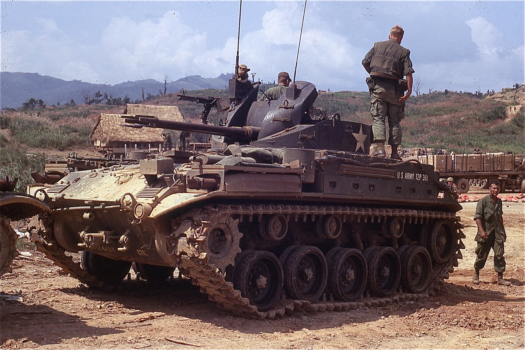 A imagem apresenta uma cena de campo militar em um ambiente tropical ou subtropical, com montanhas ao fundo sob um céu azul claro e nuvens dispersas. O foco principal é um veículo blindado americano da Segunda Guerra Mundial — especificamente, um M24 Chaffee, um tanque leve do Exército dos Estados Unidos — estacionado sobre terra batida e arenosa, possivelmente num acampamento ou base temporária.O carro está posicionado em primeiro plano, ocupando a maior parte da composição. Sua pintura é escura, provavelmente verde-oliva ou marrom escuro, típica das forças armadas americanas na época. Na lateral direita (da perspectiva do observador), há inscrições claras: “U.S ARMY 12P 381”, indicando sua pertencência à Força Armada Americana e o número de identificação. Um grande símbolo da Estrela de cinco pontas branca, característico do exército norte-americano, destaca-se no lado do casco, próximo ao canhão. Na parte superior do veículo, dois soldados estão posicionados: um sentado perto do compartimento do motorista, apoiado nas estruturas metálicas; outro, mais distante, parece estar em pé sobre o teto do tanque, olhando para frente, vestindo uniforme camuflado, colete e botas militares. Ambos parecem estar equipados com capacetes e têm aparências consistentes com os soldados da era da guerra fria ou guerras recentes após as guerras mundiais. No topo do veiculo também se vêem antenas verticais que são utilizadas como sistemas de comunicação por rádio. No centro do veículo, o canhão principal está elevado, apontando ligeiramente para cima, enquanto outra arma automática pequena — talvez um fuzil pesado ou um lançador de granadas — está instalada na posição frontal do tanque. A estrutura mecânica inclui roletes de tração robustos, rodas grandes e engrenagens visíveis nas laterais, mostrando detalhes técnicos complexos e usados intensivamente durante operações terrestres. À esquerda da tela, parcialmente oculto pelo próprio tanque, encontra-se outro veículo similar, mas menos bem iluminado, sugerindo que este pode ser apenas parte de uma frota menor de unidades de combate. Ao fundo, há alguns edifícios rústicos com telhados de palha, construídos com materiais simples, indicativos de áreas habitadas pelos civis locais, ou mesmo bases improvisadas pela força militar localizada. Um homem em pé, visto de costas, está posicionado logo atrás do tanque centralizado. Ele usa um uniforme semelhante aos outros soldados presentes e tem cabelo curto, cortado de forma moderna para aquela época. Seus braços estão cruzados, e ele parece estar observando algo fora do quadro, possivelmente coordenando atividades ou aguardando ordens. Mais longe, ainda mais ao fundo, percebe-se um grupo de soldados reunidos junto a um veículo de transporte de carga aberto, carregado com caixotes ou contêineres de madeira. Este último possui pneus largos e tracção suave, adequado para viagem em território difícil. Ao redor do cenário natural, a paisagem mostra colinas cobertas de vegetação densa, especialmente arbustos baixos e árvores esparsas, formando uma floresta misturada entre campos abertos. As cores predominantes são tons de vermelho avermelhado, castanhos e marrons naturais combinados com o cinza metalizado do tanque e o branco das nuvens no horizonte. Em geral, toda a cena transmite uma atmosfera de tensão e preparação, com elementos de realismo histórico e documental, capturando momentos cotidianos dentro de uma operação militar. Os detalhes tecnicamente precisos permitem inferir que esta fotografia foi feita em algum momento posterior às Guerras Mundiais, sendo muito possível que tenha sido realizada durante conflitos regionais ou testes de mobilidade militar nos anos 50/60. Além disso, a presença de vários indivíduos indica que houve alguma organização logística envolvendo múltiplos membros da equipe.