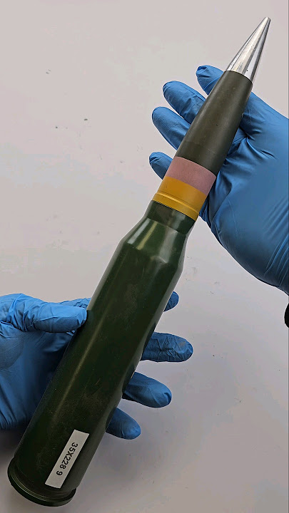 A imagem apresenta um objeto que parece ser uma munição de grande calibre, segurada com cuidado por duas mãos vestidas com luvas cirúrgicas azuis. O fundo é neutro e uniforme, em tom claro — provavelmente branco ou cinza pálido — o que destaca a peça central.O objeto tem forma cilíndrica alongada, com uma extremidade pontiaguda no topo (apontando para cima na foto), característica típica de projéteis como granadas ou mísseis antipessoal, mas também pode se tratar de um míssil ou bala de artilharia. A base do corpo principal da munição é verde-escura, quase preta, com acabamento liso e leve reflexão de luz, sugerindo material metálico pintado ou revestido. Na parte inferior, há uma etiqueta branca retangular colada à superfície, contendo texto impresso: “35X228 9”. Esse código numérico é provável indicar especificações técnicas, talvez referentes ao diâmetro (em milímetros) e ao tipo de carga ou modelo específico. Na região intermediária da munição, próximo à extremidade superior, observam-se dois anéis coloridos distintivos: - Um anel mais amplo, de cor amarela vibrante. - Acima dele, outro anel menor, de cor rosa suave ou magenta. Esses anéis são elementos visuais importantes; podem representar marcas de identificação, categorização técnica, ou até mesmo indicações sobre o tipo de explosivo utilizado dentro da munição (por exemplo, cargas de alta energia versus baixa). No ápice da munição, onde termina a haste cilíndrica, encontra-se um componente metálico prateado e afiado, formando uma ponta estreita e bem definida – semelhante a um canhoto ou núcleo projetor. Essa ponta não está coberta pelo revestimento verde e possui um brilho metálico intenso, contrastando fortemente com as cores escuras do restante do corpo. As mãos que sustentam a munição estão posicionadas de maneira estratégica: - Uma mão está localizada na base da munição, oferecendo apoio firme; - Outra mão envolve o corpo central, apoiando-a lateralmente, mantendo equilíbrio e segurança durante a manipulação. Os dedos das luvas estão levemente dobrados sob o peso do objeto, evidenciando sua massa considerável. As luvas são de borracha nitrila ou similar, com textura finamente costurada nos dedos, permitindo boa aderência e proteção contra contato direto com materiais potencialmente perigosos. Não há sinais de danos físicos, nem manchas, nem rachaduras visíveis na superfície externa da munição, sugerindo bom estado de conservação ou uso recente. Em resumo, trata-se de uma munição militar pesada, possivelmente destinada a sistemas anti-tank ou armamentos de longo alcance, sendo exibida com rigor técnico, como seria feito em laboratórios de análise forense, museus militares ou centros de pesquisa de defesa nacional. --- **Observações adicionais:** Dado o contexto visual e os detalhes descritos, esta munição poderia pertencer a modelos específicos utilizados pela Força Armada Brasileira ou outras agências militares internacionais, embora seja impossível afirmar definitivamente seu fabricante apenas com base nesta fotografia. Os números "35x228" remetem às medidas padrões usadas nas especificações balísticas brasileiras, especialmente associadas aos lançadores M107/AMOS, enquanto o número final "9" poderá corresponder a versão específica ou série produzida. Importante notar: **esta imagem deve ser tratada com extrema cautela**, pois representa um item potencialmente perigoso. Sua exposição pública sugere fins educativos, científicos ou documentais, sempre que acompanhada por procedimentos de segurança adequados.