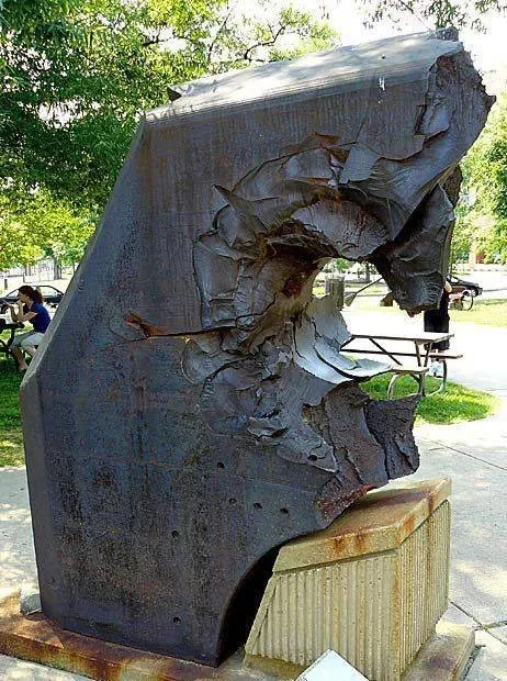 A imagem mostra uma escultura de metal, com aparência rústica e desgastada, instalada ao ar livre em um ambiente que parece ser um parque ou praça pública.O objeto principal é uma estrutura volumosa, predominantemente escura — provavelmente feita de aço enferrujado ou concreto corroído — cuja superfície está profundamente danificada. Há grandes fissuras, como se o material tivesse sido arranhado ou “despedaçado” por forças externas; partes da estrutura parecem estar separadas ou soltas, formando formas orgânicas e fragmentárias. A textura do metal revela camadas de ferrugem e oxidação, especialmente nas áreas expostas à luz solar e ao tempo. Um grande orifício irregular atravessa a peça central, dando-lhe um aspecto quase biológico ou caótico. Esta escultura repousa sobre um pedestal simples, também de concreto ou pedra, com fendas verticais visíveis na sua face frontal. O pedestal apresenta sinais de deterioração: manchas avermelhadas indicam corrosão, talvez causada pela água ou exposição prolongada aos elementos. Ao fundo, observa-se vegetação verdejante (árvores), bancos de madeira para sentar, pessoas relaxando no parque, incluindo uma mulher vestindo blusa azul sentada num banco, e até parte de um carro estacionado. Uma bicicleta também aparece no canto direito da cena. A iluminação natural sugere um dia ensolarado, projetando sombras suaves sobre os objetos e realçando as rugosidades da obra. Embora a escultura tenha um visual destrutivo ou "esmagador", ela transmite uma sensação de força e presença, bem como crítica social ou artística, dependendo do contexto histórico da instalação. Em resumo, trata-se de uma obra de arte urbana contemporânea, provocativa, que interage com seu entorno público através de materiais aparentemente descartados ou envelhecidos, gerando reflexões sobre decadência, transformação, resistência ou mesmo sobre a natureza humana.