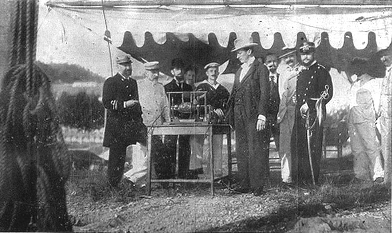 Esta é uma fotografia em preto e branco, com aparência antiga e de alta qualidade documental, capturando um momento histórico ao ar livre sob uma tenda grande com bordas onduladas, típica de eventos públicos ou exposições do século XIX ou início do XX.A cena ocorre num terreno irregular coberto por grama seca e terra, sugerindo que o evento aconteceu em um local aberto — talvez um campo, parque ou área de exibição temporária. Ao fundo, percebe-se uma paisagem natural composta por árvores e colinas suaves, indicando que a foto foi tirada em um ambiente rural ou semi-rural. No centro da composição está uma mesa simples de madeira, apoiada sobre quatro pernas retangulares. Sobre ela repousam dois objetos principais: à esquerda, parece ser um modelo mecânico complexo, possivelmente um pequeno motor, uma máquina de vapor ou até mesmo um protótipo de veículo; ele possui estruturas metálicas visíveis e componentes que parecem estar conectados por tubulações. À direita deste objeto, há outro dispositivo menor, também montado sobre uma base, cuja função não é clara pela imagem mas pode ser um instrumento científico ou técnico. Ao redor desta mesa estão reunidos aproximadamente dez homens, vestidos com roupas formais da época: - **À esquerda**, um homem alto e robusto veste um uniforme militar formal escuro (possivelmente naval), com galões nos braços e botas altas; seu olhar está voltado para os outros participantes. - Próximo a ele, um segundo homem usa um casaco claro de estilo europeu da época, com gola e punhos bem definidos; tem barba e cabelos curtos, e parece estar observando atentamente o equipamento na mesa. - Entre eles, mais atrás, outra figura masculina aparece com traje elegante, inclinando-se ligeiramente sobre a mesa como se estivesse examinando algo próximo aos dispositivos expostos. - No lado oposto, um homem de terno longo e chapéu fedora (ou boina) destaca-se pelo seu porte ereto e postura confiante, enquanto conversa com alguém perto dele. Ele parece ser o foco central da atenção dos presentes. - Mais à direita, um oficial militar com uniforme escuro adornado com botões dourados e medalhas no peito, além de uma espada presa à cintura, demonstra autoridade. Seu capacete é característico das forças armadas nacionais daquela era. - Outros indivíduos completam o grupo: alguns usam ternos civis, outras camisas sociais sem gravata, e ainda há mulheres entre os espectadores – duas delas são particularmente notáveis: uma jovem com roupa social leve, capuz e blusa justa, e outra mulher mais velha usando um vestido tradicional, ambas observando as apresentações com interesse. O cenário geral transmite uma atmosfera de prestígio, cerimônia pública ou exposição científica/tecnológica. As expressões faciais variam desde o entusiasmo ao respeito profundo, refletindo a importância do evento. A luz solar incide diretamente sobre os personagens, criando sombras claras nas laterais, evidenciando que a sessão fotograficamente foi realizada durante o dia. Os detalhes finos — como as texturas das roupas, os padrões dos sapatos, os ornamentos militares, e inclusive o desgaste perceptível da própria tela da tenda — reforçam a autenticidade histórica da imagem. Embora seja difícil identificar exatamente quem são essas pessoas ou qual o contexto específico dessa exposição, a fotografia oferece uma janela fascinante para entender a cultura tecnológica e institucional da Europa ocidental no final do século XIX ou começo do século XX.