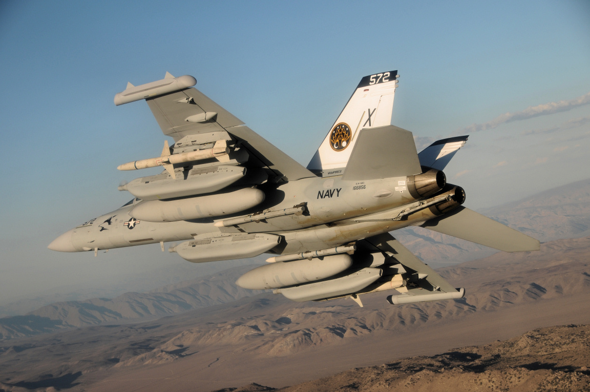A imagem apresenta um avião de combate da Marinha dos Estados Unidos, especificamente um F/A-18 Hornet (ou possivelmente uma variante como o F/A-18E/F Super Hornet), em voo sobre um território árido e montanhoso sob um céu azul claro com poucas nuvens.**Detalhes do Avião:** * **Código Identificador:** Na cauda vertical, destaca-se claramente a designação "572" em letras pretas no topo, abaixo da qual está a letra "X", indicando que se trata de um avião pertencente à unidade VFA-140 ou similar — embora os códigos específicos podem variar conforme as operações. * **Símbolo da Marinha dos EUA:** A fuselagem exibe a palavra “NAVY” pintada em preto na parte inferior da secção central, logo acima das turbinas dos motores. Também é visível o símbolo estrelado da Marinha nos flancos, próximo ao nariz. * **Emblema da Unidade / Grupo Operacional**: No centro da cauda, há um distintivo circular dourado com elementos gráficos complexos, provavelmente representando o emblema de uma unidade específica dentro da Força Aérea Naval. O padrão parece ser inspirado num grupo de combatentes ou escolta naval. * **Configuração Armamentária:** O avião está equipado com diversos sistemas de armamento: * Um sistema de mísseis guiados por radar instalado nas ponteiras laterais das asas; * Uma bomba tipo M61 para bombardeiros estruturais posicionadas sob cada asa; * Cilindros cilíndricos grandes pendurados debaixo do corpo principal – esses são provavelmente mísseis anti-navio ou granadas lançáveis de longo alcance; * Outras placas suspensórias menores estão fixadas aos lados inferiores das asas, sugerindo capacidades adicionais de ataque. * **Asas Estendidas/Reconfiguradas:** As asas têm forma típica de jato moderno, mas parecem estar configuradas numa posição mais baixa durante o voo, talvez preparando-se para manobrar ou ajustar velocidade aerodinâmica. * **Motores Ejetivos:** Os dois motores turbofan estão integrados às extremidades traseiras do fuselagem; seus tubos de escape são largos e robustos, característicos de aviões militares de alto desempenho. * **Pintura e Corpo Principal:** O avião tem cor cinza-tanque uniformemente aplicada, típico de configurações de camuflagem militar contra fundos terrestres claros. **Contexto Visual:** O avião voa bem acima do solo, oferecendo uma vista panorâmica ampla do cenário abaixo: * **Terreno Terrestre:** Abaixo do avião, observam-se extensos desertos e colinas rochosas com tons predominantemente marrom-acastanhados e bege escuro. As linhas ondulantes formadas pelas elevações geográficas criam texturas rústicas e dramáticas. * **Horizonte e Luz Natural:** O céu apresenta tonalidades suaves de azul pálido até levemente rosado próximos ao horizonte, sinalizando que pode ser amanhecer ou entardecer. A luz solar incide diretamente sobre o avião, destacando suas formas angulares e sombras projetadas sobre o chão. * **Composição Fotográfica:** A fotografia foi tirada de ângulo lateral superior, permitindo ver tanto detalhes técnicos do avião quanto sua relação espacial com o ambiente natural circundante. Há leve movimento dinâmico capturado pela câmera, transmitindo sensação de potência e agilidade. Esta cena evoca imagens de poder militar contemporâneo, onde tecnologia sofisticada opera em ambientes hostis, combinando precisão técnica com habilidade humana em condições adversas. É uma representação vívida da força aérea norte-americana em exercício realista e estratégico.