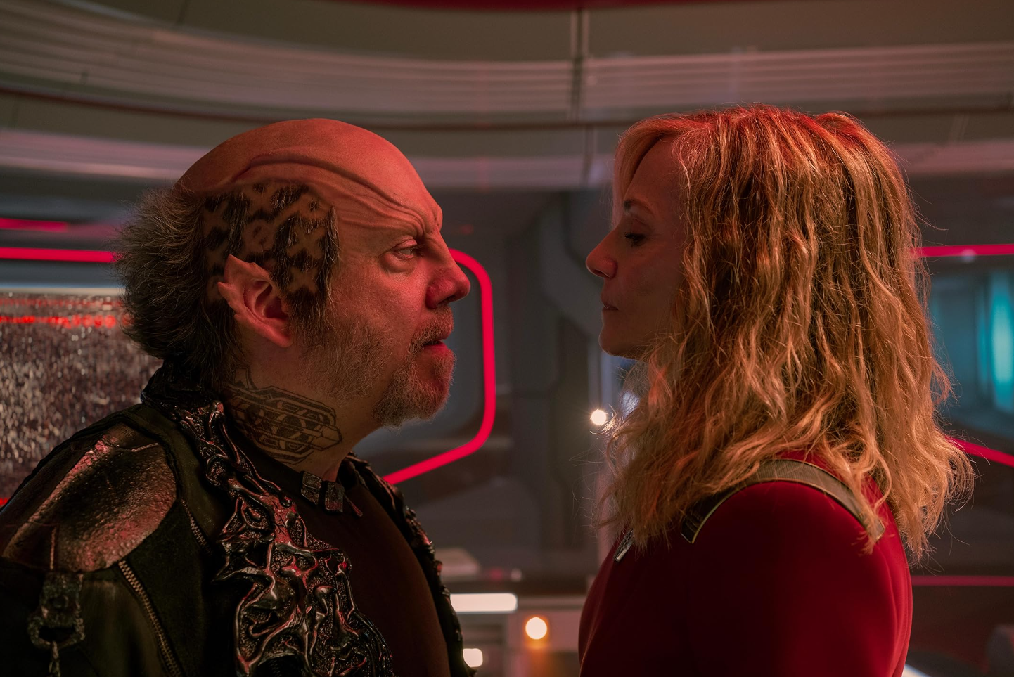 A imagem mostra dois personagens em uma cena de confronto ou tensão intensa, dentro do que parece ser um ambiente futurista e tecnológico — provavelmente a cabine de comando de uma nave espacial.À esquerda, há um homem com aparência alienígena: ele possui uma cabeça grande e redonda, pele clara e textura irregular na testa, além de cabelos escuros e encaracolados nas laterais da cabeça. Sua face é marcada por uma expressão feroz, com os olhos semicerrados e o rosto contraindo como se estivesse gritando ou prestes a atacar. Ele tem barba rala e tatuagem complexa no pescoço e na parte lateral da cabeça, parecendo feita de padrões geométricos ou tribais. Veste uma jaqueta preta com detalhes metálicos brilhantes, incluindo zíperes decorativos e adornamentos que lembram correntes ou estruturas orgânicas. À direita, está uma mulher loira, com cabelos longos, ondulados e levemente desgrenhados, iluminados pelo vermelho intenso das luzes ao fundo. Ela veste uma roupa vermelha com mangas curtas e colarinho militarizado; parece ser uniforme de oficial (talvez da frota Starfleet). Seu rosto está voltado para o homem, mas ela não demonstra medo nem raiva – sua expressão é calma, séria e determinada, quase impassível, sugerindo que ela controla a situação apesar da pressão. O cenário envolve painéis curvos, luminosos e tubulações industriais típicas de ficção científica, tudo banhado numa atmosfera avermelhada e dramática, criando um forte contraste entre as duas figuras. A composição fotográfica foca nos rostos dos personagens, destacando a tensão emocional e psicológica entre eles.