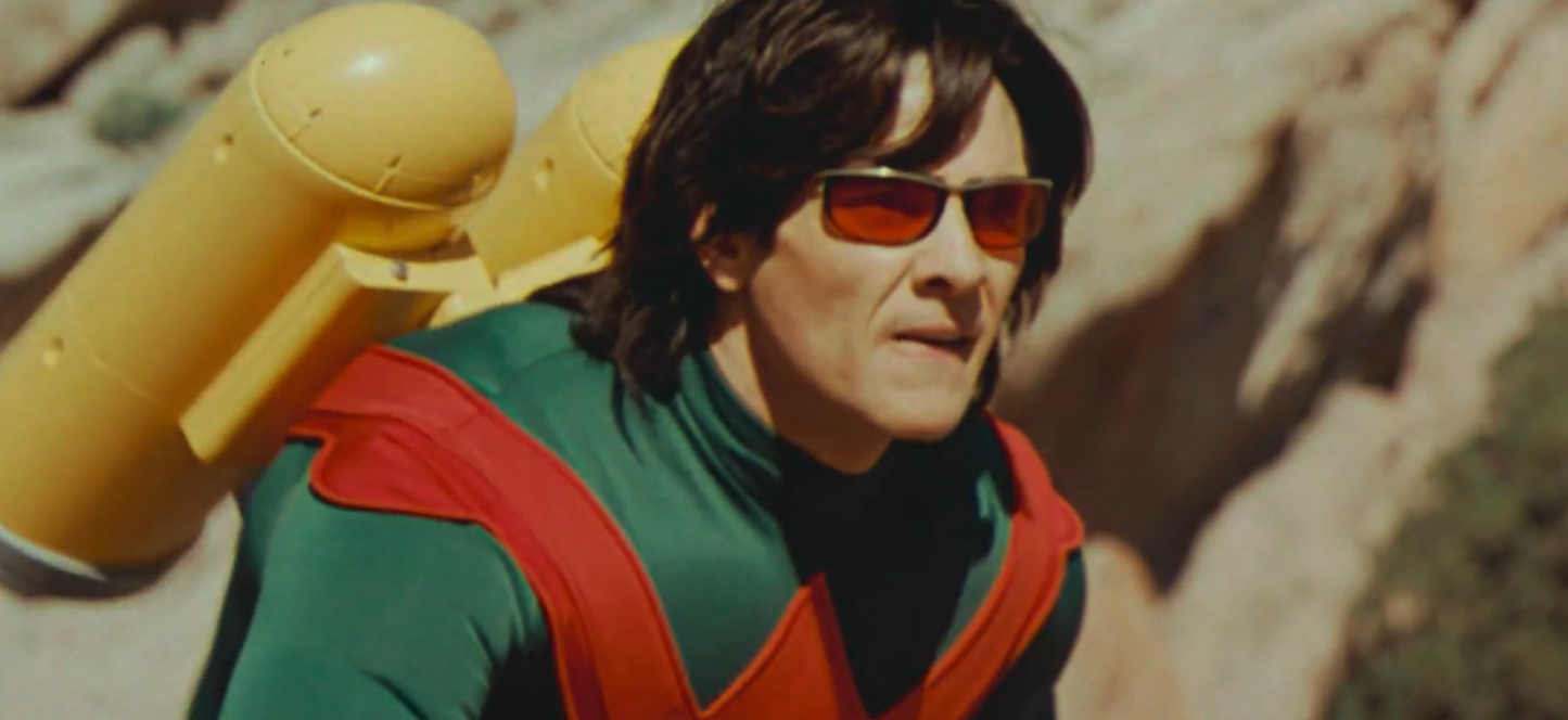 A imagem mostra um homem de meia-idade com cabelo escuro, volumoso e levemente desgrenhado, usando óculos de sol com lentes vermelhas e armação metálica fina. Ele está vestindo uma roupa que parece ser um traje ou uniforme: uma camisa verde-escura com detalhes em vermelho vibrante nas mangas e no peito, sobre uma peça preta por baixo.Sobre os ombros dele há dois grandes cilindros amarelos, parecendo equipamentos futuristas ou tecnológicos — possivelmente armazenadores de energia ou propulsores — fixados a um suporte também amarelo. O design dos objetos é robusto, com superfícies lisas e formas arredondadas na parte superior. O fundo da cena é uma paisagem árida e rochosa, provavelmente um deserto ou montanha, com tons terrosos e sombras profundas criadas pela luz solar direta. A iluminação sugere um dia ensolarado intenso, projetando contrastes fortes entre as áreas claras e escuras do cenário. O rosto do homem tem expressão séria ou concentrada; ele olha para fora do quadro à sua direita (à esquerda do espectador), como se estivesse observando algo ao longe. Sua boca está leve aberta, indicando movimento ou atenção atenta. O estilo visual da imagem é cinematográfico, com cores saturadas e uma qualidade de filme que pode sugerir uma produção de ficção científica ou drama épico. Não há texturas muito nítidas nos fundos, mas sim um foco claro no personagem central.
