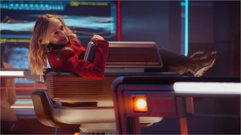 A imagem mostra uma cena de ficção científica, provavelmente dentro de um cockpit ou centro de controle de nave espacial futurista.No centro da composição está uma jovem mulher com cabelos loiros ondulados e compridos, que se encontra relaxada em uma cadeira ergonômica e moderna. Ela veste um uniforme vermelho vibrante, típico de personagens militares ou cientistas de exploração espacial, com detalhes metálicos nas mangas e no peito do casaco. Seus pés estão descalços sobre o console, calçando botas altas escuras, sugerindo um ambiente funcional e tecnológico. Sua expressão é serena e pensativa; ela olha para a câmera com um leve sorriso, transmitindo confiança e tranquilidade apesar do cenário dinâmico ao redor. O fundo apresenta painéis luminosos azuis e ciano exibindo gráficos complexos e dados, indicativos de sistemas de navegação, comunicação ou monitoramento. A iluminação geral é quase totalmente artificial — tons frios predominam nos telões, contrastando com os reflexos dourados e laranjas das luzes internas dos consoles e dispositivos próximos à protagonista. Um destaque visual importante é um pequeno LED quadrado emitindo luz amarela intensa na parte inferior direita da imagem, próximo a um elemento estrutural angular do equipamento. A estética da sala combina elementos retrofuturísticos: linhas curvas suaves combinadas com superfícies lisas e materiais como plástico brilhante e metal polido. O design evoca décadas passadas (como os anos 70-80) mas reinterpretado num contexto científico avançado, reforçando a atmosfera de aventura espacial. Em resumo, a foto transmite uma sensação de elegância técnica, conforto operacional e personalidade forte, retratando alguém dominando seu espaço enquanto descansa — talvez durante uma pausa estratégica antes de enfrentar novos desafios intergaláticos.