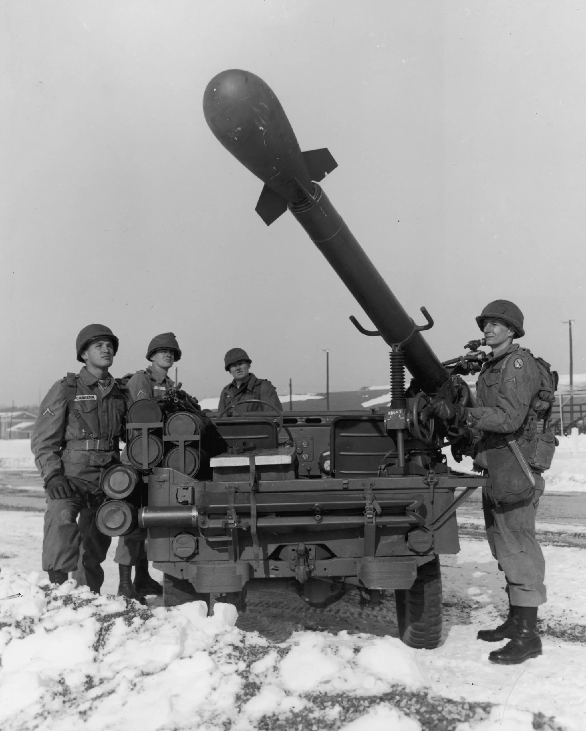 Imagem histórica em preto e branco de quatro soldados do Exército dos Estados Unidos posando ao lado de um lançador de míssil tático nuclear Davy Crockett durante a Guerra Fria. O míssil, em forma de projétil alongado com ogiva esférica na ponta, está montado em uma plataforma móvel inclinada para cima, com canos de lançamento múltiplos e estrutura metálica visível. Os soldados usam uniformes de combate, capacetes e botas pesadas; um deles está segurando o mecanismo de disparo. Ao fundo, neve cobre o solo e há construções militares ao longe. A foto transmite a tensão da era nuclear, com foco na arma tática de curto alcance projetada para uso em campo de batalha.