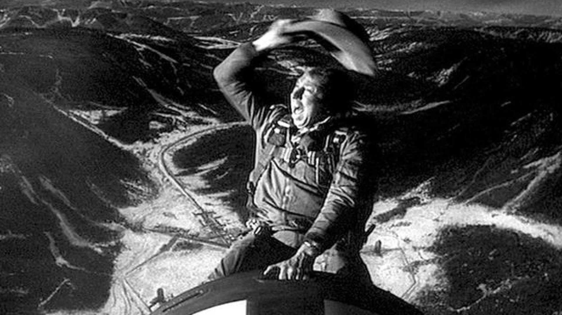 Imagem em preto e branco de um cowboy de chapéu largo, montado em uma bomba nuclear, com uma expressão de espanto ou admiração. Ele está com o braço direito levantado, a mão na aba do chapéu como se estivesse protegendo os olhos do sol ou do vento, olhando para o horizonte. Ao fundo, uma vasta paisagem desértica dramática com montanhas escarpadas, vales profundos e estradas sinuosas cortando o terreno árido. A composição cria um contraste marcante entre a figura humana solitária e a imensidão da natureza selvagem, transmitindo sensação de aventura, descoberta e escala épica.