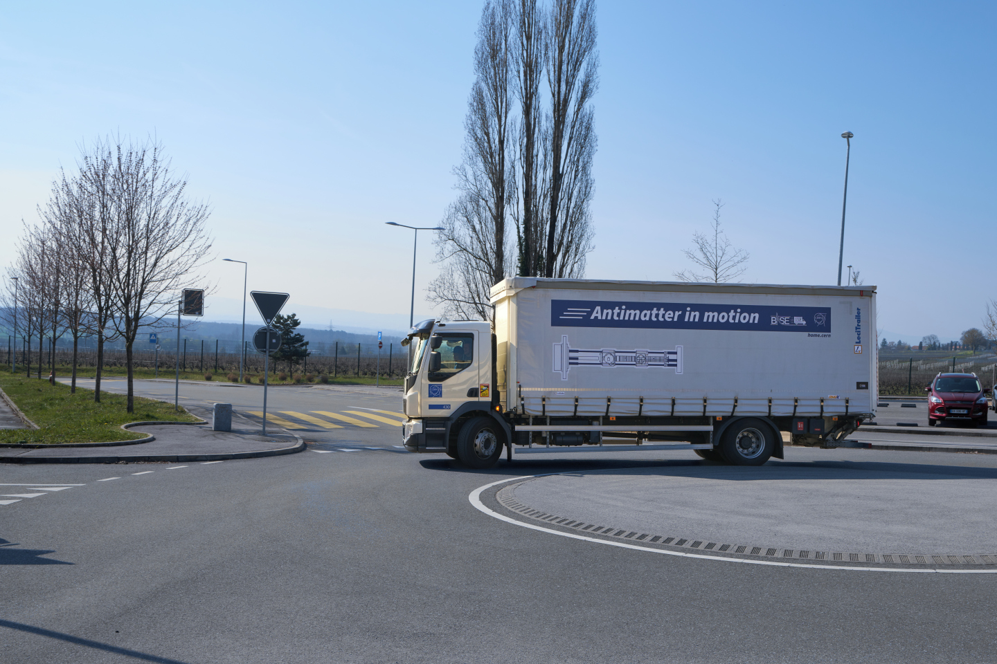 A imagem mostra um grande caminhão branco com as palavras “Antimatter in motion” escritas na lateral. O caminhão circula em uma rotatória em uma estrada com céu azul ao fundo. Há árvores do lado esquerdo da estrada e um carro vermelho do lado direito. Ao longe, há montanhas e um céu azul claro. A estrada parece estar vazia, sem carros ou pessoas visíveis.