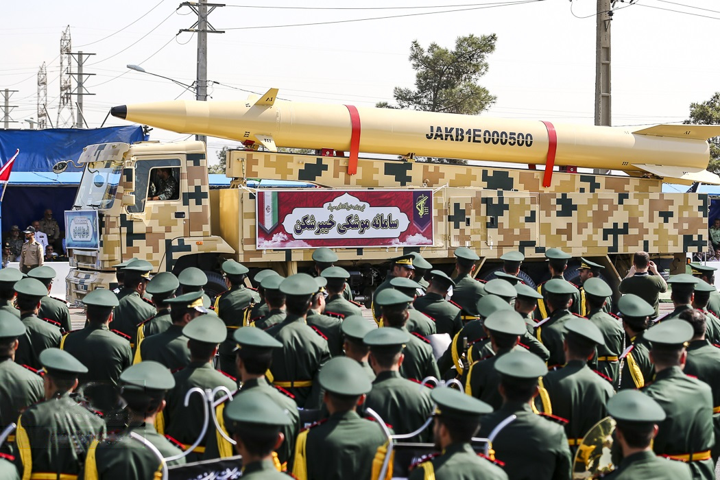 A imagem mostra um desfile militar com um grande caminhão carregando um grande míssil. O caminhão é decorado com um padrão camuflado e tem um banner na lateral que diz "Jammu e Caxemira". O míssil é amarelo e vermelho e está montado no topo do caminhão. O desfile acontece em uma rua com linhas de energia e árvores ao fundo.Em primeiro plano, há um grande grupo de soldados vestindo uniformes verdes e chapéus verdes, marchando em formação. Eles seguram faixas e bandeiras, e alguns deles seguram instrumentos musicais. O céu está azul e o tempo parece ensolarado e claro.