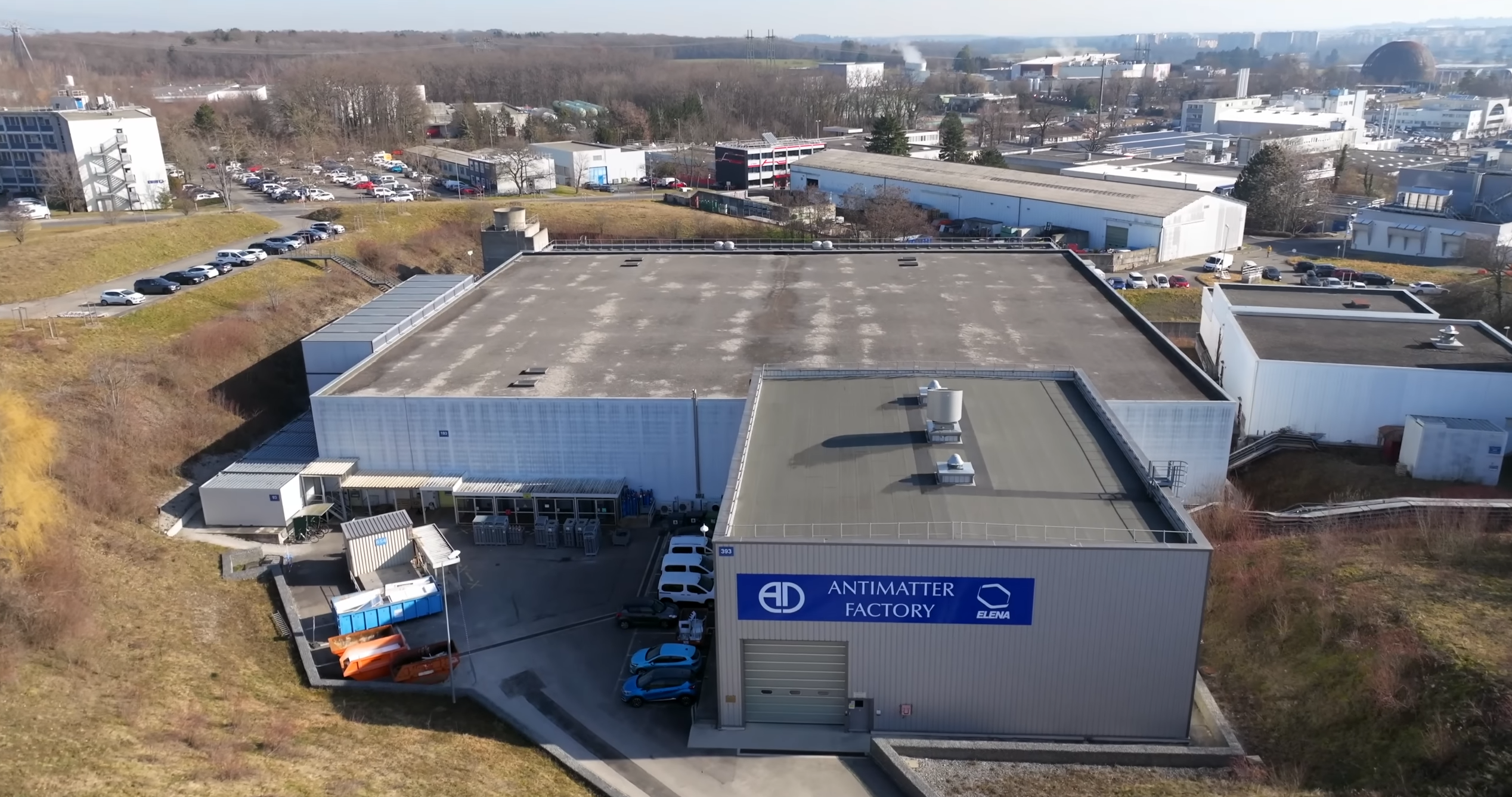 A imagem mostra uma vista aérea de um grande edifício industrial com telhado plano. O prédio é cinza e tem uma placa azul que diz “Fábrica de Antimatéria”. Existem vários carros estacionados em frente ao prédio e alguns outros edifícios e estruturas ao fundo. O céu está azul e há árvores e colinas ao longe. A imagem parece ter sido tirada de um ponto elevado, olhando para o edifício.