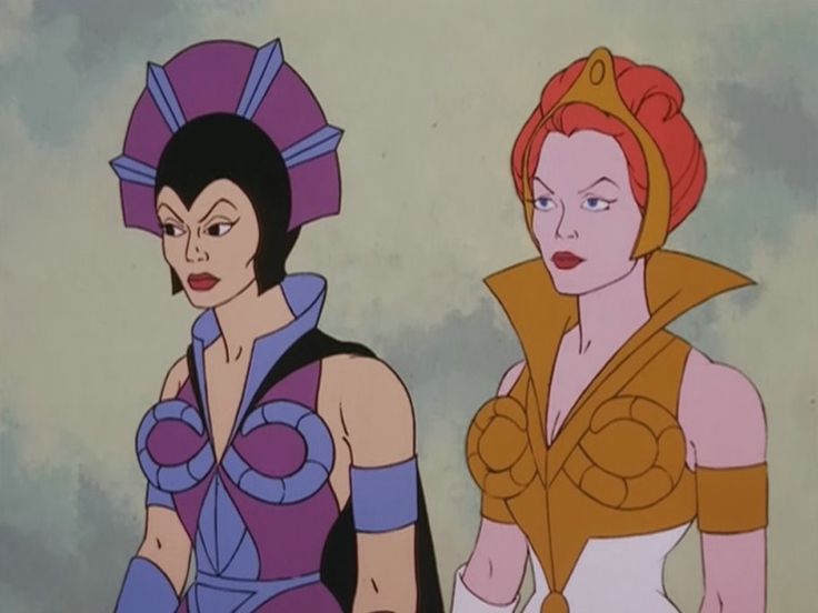 Esta é uma captura de tela de uma cena de desenho animado, provavelmente da série "He-Man e as Forças do Mal".**Visão geral:** A imagem exibe um plano médio de duas personagens femininas em pé, lado a lado, com o fundo desfocado em tons suaves de verde e cinza. O estilo visual é característico da animação ocidental dos anos 80, com traços definidos, cores planas e uma paleta de cores vibrante. **Elementos principais:** Dois personagens estão em destaque: 1. **Personagem à esquerda:** Possui cabelo preto curto e liso. Ela usa um traje predominantemente roxo e azul, com detalhes em preto. Seu design inclui uma peça de cabeça ou tiara roxa com pontas azuis e uma armadura com padrões circulares nos ombros. Ela tem uma faixa roxa no braço esquerdo. 2. **Personagem à direita:** Tem cabelo vermelho preso em um coque alto, coberto por uma tiara dourada. Seu traje é composto por tons de dourado, branco e laranja, com um colarinho alto e drapeado. Ela possui uma armadura dourada com texturas de círculos entrelaçados nos ombros e uma faixa dourada no braço esquerdo. **Detalhes visuais relevantes:** Ambas as personagens têm rostos maquilhados com delineador de olhos preto e batom vermelho. A iluminação é suave e uniforme, típica da animação clássica, sem sombras fortes. A composição é simples, focando nos rostos e no design dos trajes, que parecem ser uniformes de batalha ou mágicos. **Texto presente:** Não há texto visível na imagem. **Estilo e atmosfera:** A atmosfera é de ação ou confronto, sugerida pelas expressões sérias e pela postura das personagens. O estilo visual é distinto de uma animação moderna, remetendo à estética dos anos 80.