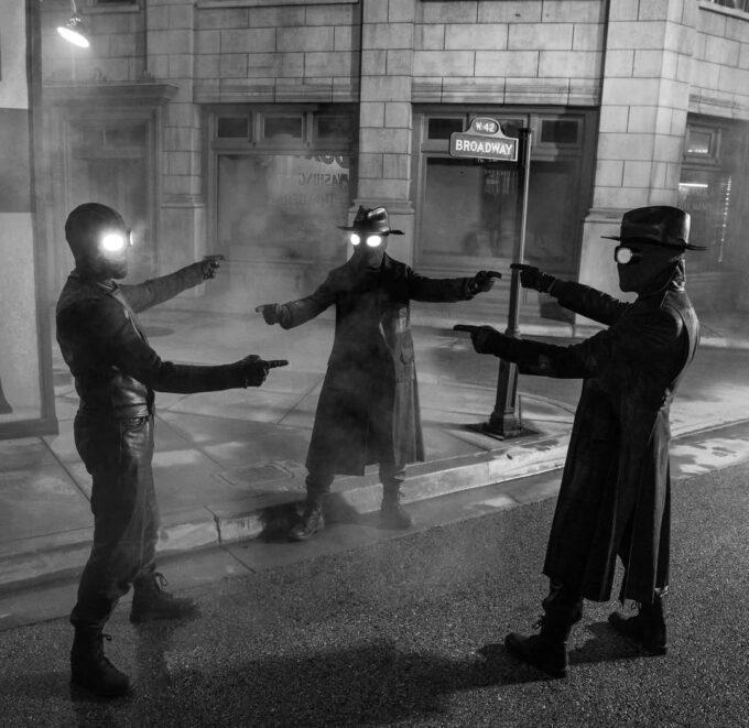 Esta fotografia em preto e branco retrata uma cena de confronto urbano noturno, caracterizada por uma atmosfera densa de neblina ou fumaça. Três indivíduos, posicionados em formação triangular, ocupam o centro da composição, com dois deles à esquerda e direita e um terceiro no centro, ligeiramente mais distante. Todos vestem casacos longos e chapéus de aba larga, e usam máscaras faciais que cobrem o rosto inteiro, exceto por duas lentes brilhantes e redondas que simulam olhos, emitindo uma luz intensa e focalizada. Cada figura segura uma arma de fogo apontada para o centro do grupo, criando uma tensão visual imediata. O cenário é um quarteirão de cidade com calçada e rua visíveis, onde um poste de sinalização de rua exibe claramente as inscrições "W 42" e "BROADWAY". A iluminação provém das próprias lentes dos personagens e de uma fonte artificial elevada à esquerda, projetando sombras duras no chão molhado ou úmido. A textura da imagem é granulada, típica de fotografia de filme antigo ou estilização de alto contraste, reforçando o clima noir e cinematográfico. A câmera está posicionada em nível dos olhos, com enquadramento que inclui as figuras até a parte inferior do corpo, destacando a simetria e a ação dinâmica do momento.