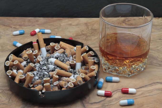 Foto de uma mesa de madeira, e sobre ela há um copo de whisky abaixo da metade, um cinzeiro cheio de bitucas de cigarro, e pílulas espalhadas (Crédito: Luis Angel Garcia/Adobe Stock)