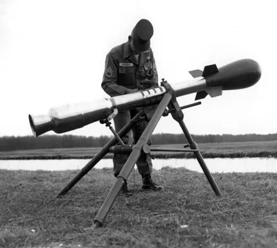 Esta é uma fotografia em preto e branco de um homem em uniforme militar ajustando uma arma de lançamento de projétil montada sobre um tripé. O sujeito, que aparenta ser adulto, usa boné militar, camisa de manga comprida com insígnias nos ombros e calça combinando com botas. Ele está de pé, inclinado levemente para frente, com as mãos na parte frontal do lançador, concentrado em uma peça de ajuste ou verificação. A arma é longa, cilíndrica, com uma ponta metálica brilhante e cauda estabilizadora com estabilizadores triangulares. O tripé é robusto, de três pernas, apoiado em terreno gramado. Ao fundo, há uma faixa de água horizontal e uma linha de árvores distantes, sugerindo um campo aberto ou área de treinamento. A iluminação é difusa, provável de céu nublado, criando sombras suaves e sem contraste forte. A câmera está em nível médio, capturando o sujeito e a arma em plano inteiro, com foco no equipamento e na postura do operador. Não há texto visível na imagem. O estilo é documental, típico de arquivo histórico militar, com composição centralizada e ênfase na precisão técnica da cena.