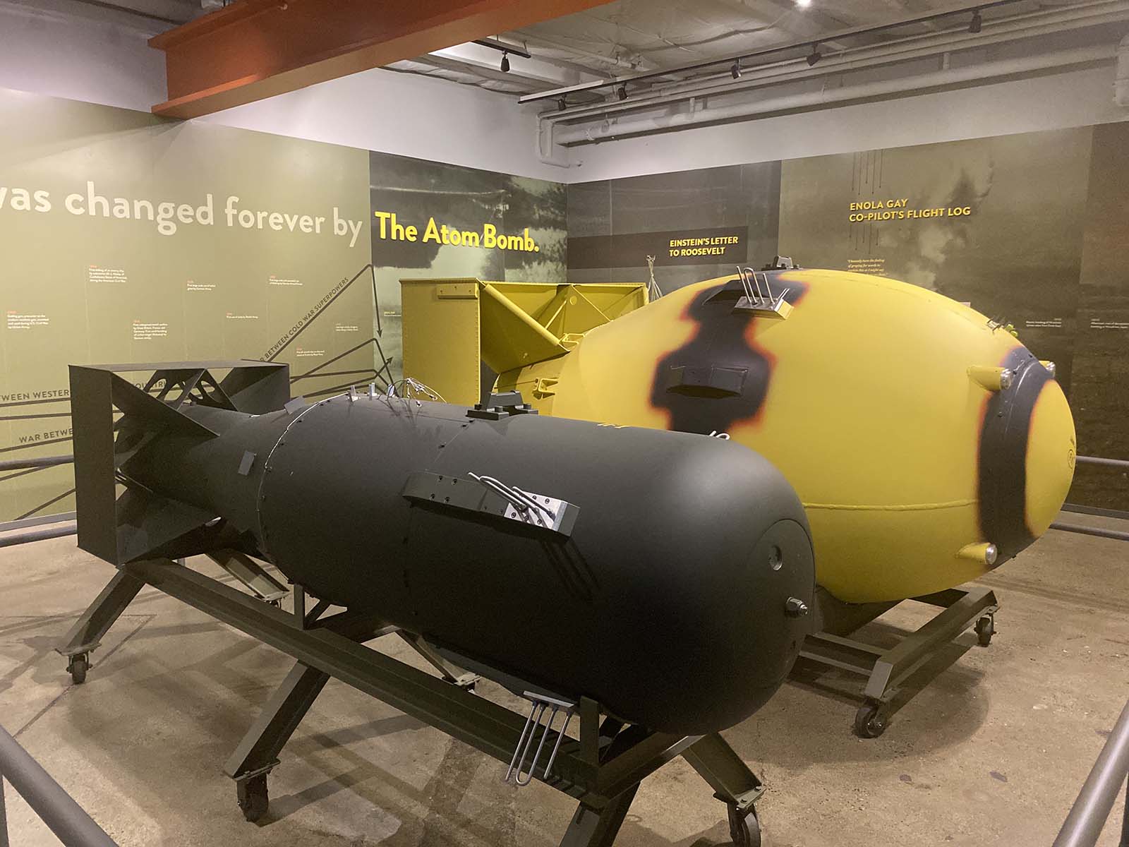 Em um ambiente interno de museu, com iluminação artificial focada e teto industrial visível, duas bombas atômicas em tamanho real são exibidas sobre suportes metálicos com rodas. A bomba mais próxima, em primeiro plano, é preta fosca, com acabamento metálico liso e detalhes como parafusos, plaquetas de identificação e pequenos alçaques de aço. Sua forma é alongada, com um nariz arredondado e estabilizadores triangulares na cauda. Logo atrás, posicionada em segundo plano, está a bomba amarela, coberta por uma pintura vibrante com uma mancha escura simulando queimadura no centro, e detalhes em preto ao redor das abas e fixações. Ambas as bombas repousam sobre estruturas de aço pintado em verde-oliva, com trilhos de rolamento e apoios angulares. O piso é de concreto cinza claro, com reflexos suaves das luzes do teto, que são spotlights direcionados para os objetos, criando sombras definidas sob os suportes. No fundo, painéis informativos na parede em tons de verde-oliva e preto exibem textos em branco e amarelo, incluindo frases como "The Atom Bomb", "EINSTEIN'S LETTER TO ROOSEVELT" e "ENOLA GAY CO-PILOT'S FLIGHT LOG". A composição é equilibrada, com as bombas alinhadas em diagonal leve, guiando o olhar do espectador da frente para trás. A profundidade de campo é moderada, mantendo ambas as bombas nítidas, enquanto o fundo permanece levemente suave. A atmosfera é sóbria e documental, típica de exposição histórica, com foco na materialidade dos objetos e na informação textual complementar.